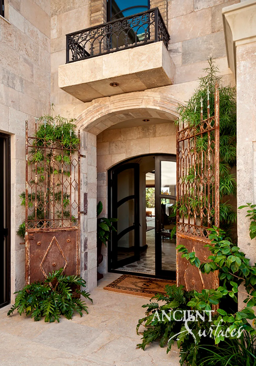 Arched antique limestone entrance framed by reclaimed iron gates, with coursed limestone wall cladding and a carved stone balcony above