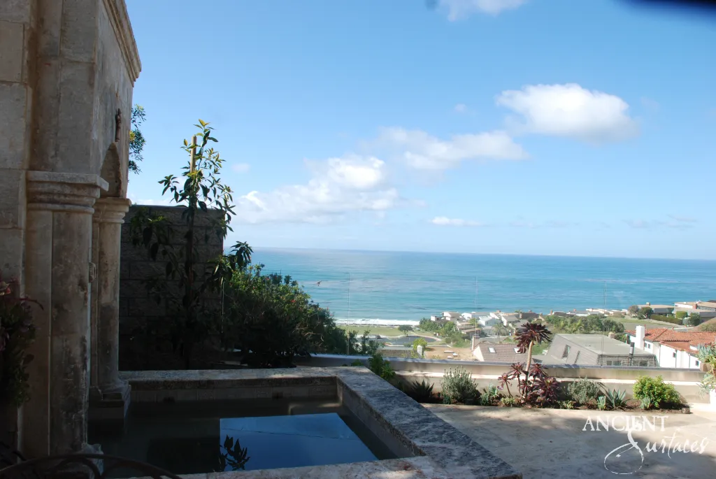 Carved limestone wall fountain standing over a terrace basin coped in reclaimed stone, looking out across the ocean