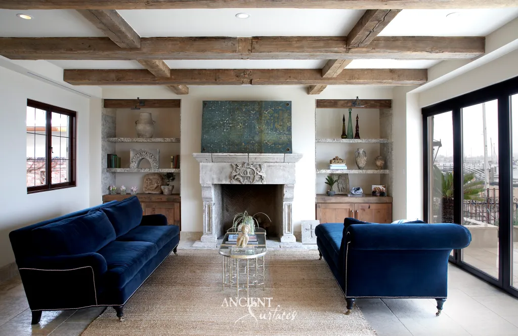 Carved limestone fireplace mantel centered on a floral cartouche, set between built-in shelves beneath old timber beams