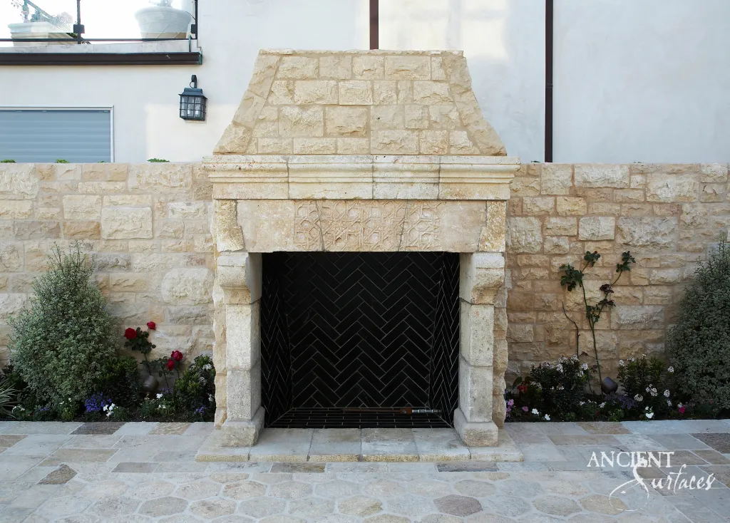 Outdoor limestone fireplace mantel with a carved frieze and tapered stone hood, built into a limestone garden wall