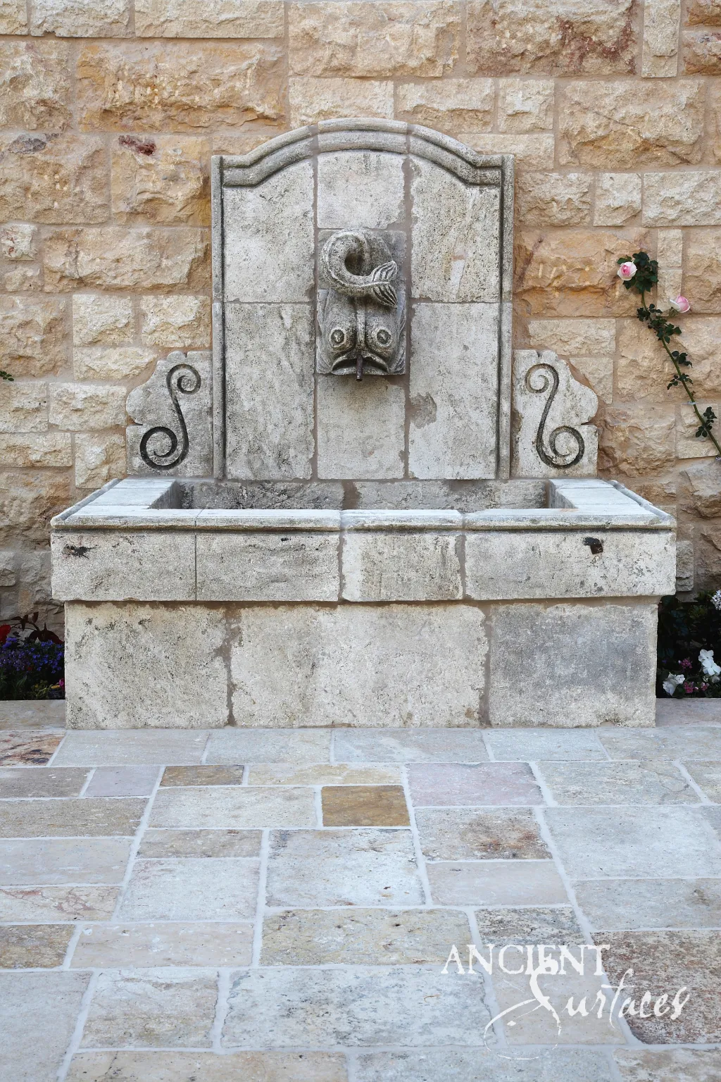 Antique limestone wall fountain with a carved fish spout and scrolled wings above a solid stone trough basin