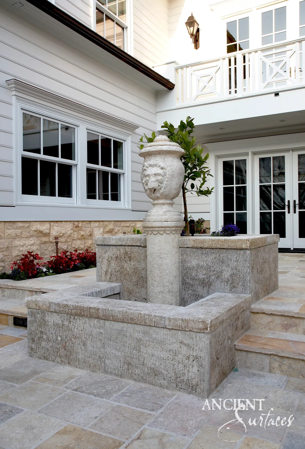 Square limestone fountain basin gathered around a central column topped by a carved lion head urn, set into limestone paving